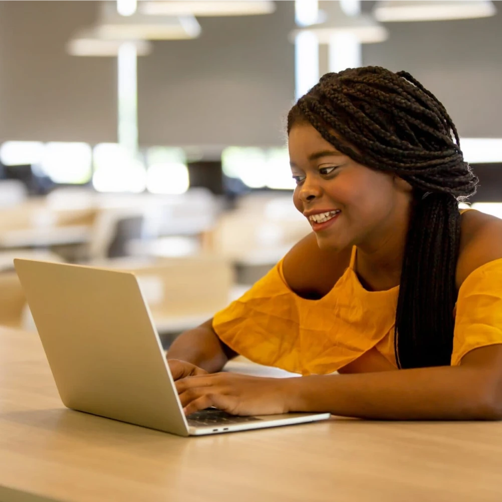 Smiling woman in yellow shirt working on laptop at desk in bright modern office.