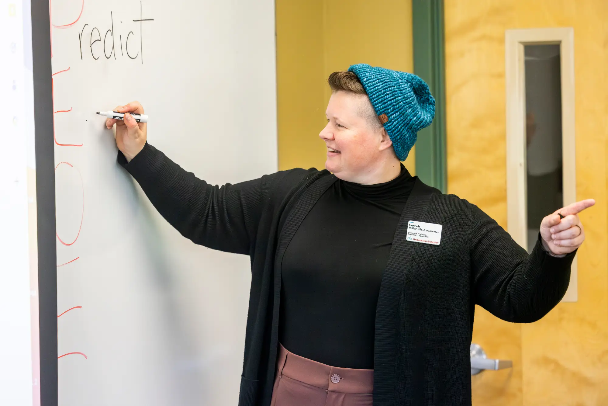 A smiling instructor in a blue knit hat gestures toward a whiteboard while writing the word “predict” during a classroom lesson.