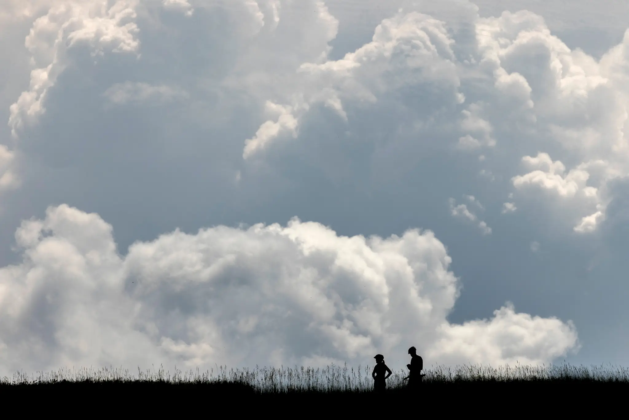 Two silhouetted figures stand in a grassy field against a dramatic sky filled with billowing white and gray clouds.