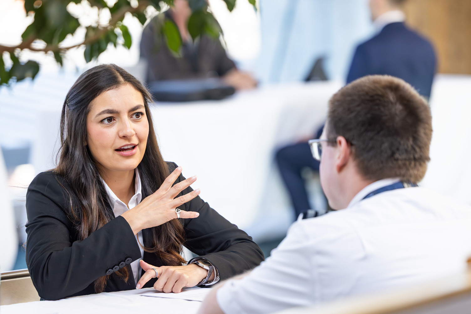 A woman in a black suit is sitting at a table and gesturing during a conversation. A man is sitting across from her with his back to the camera.