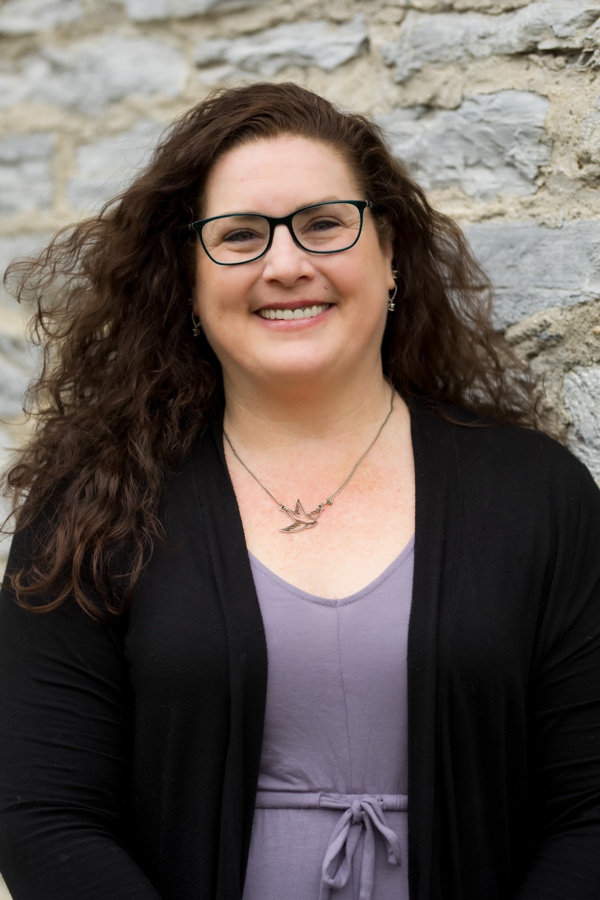 Shanti McKenna, a woman with long brown curly hair, is wearing a purple shirt with a black cardigan. She's standing outdoors and smiling at the camera.