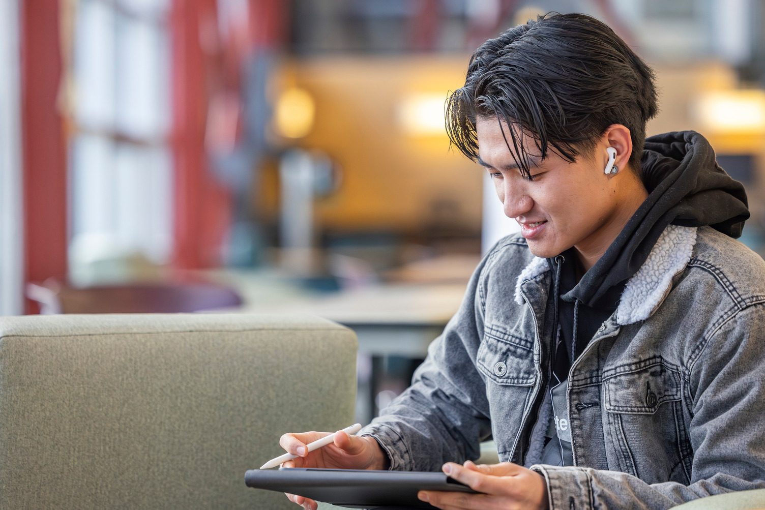 A young man sitting in a well-lit lobby, looking at an ipad in his hands with headphones in.