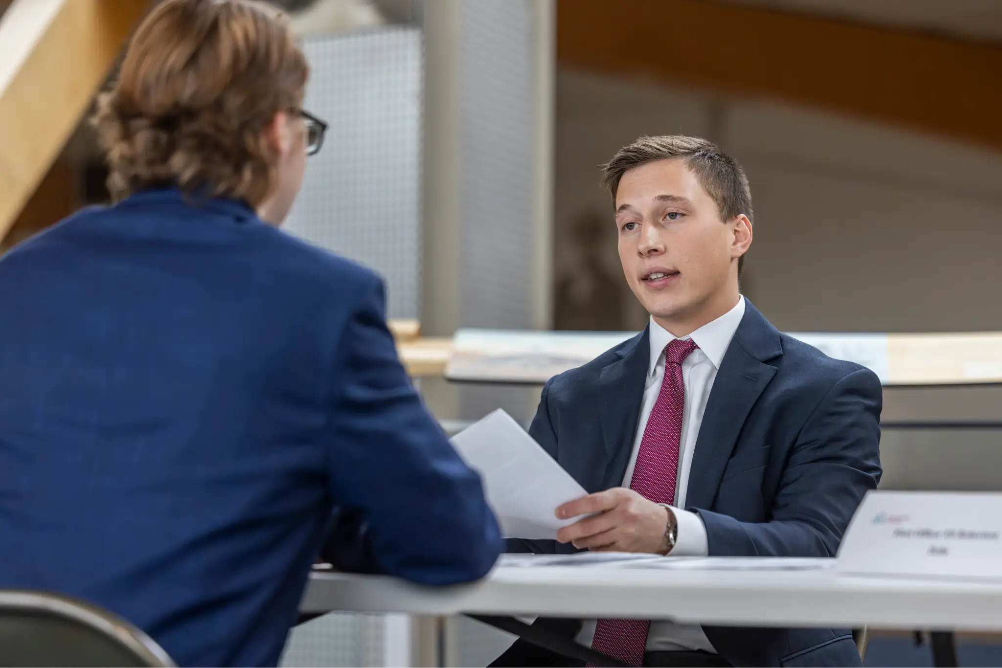Two men in suits are sitting at a table together. One is holding a stack of papers in his hands, while the man in the foreground has his back to the camera.