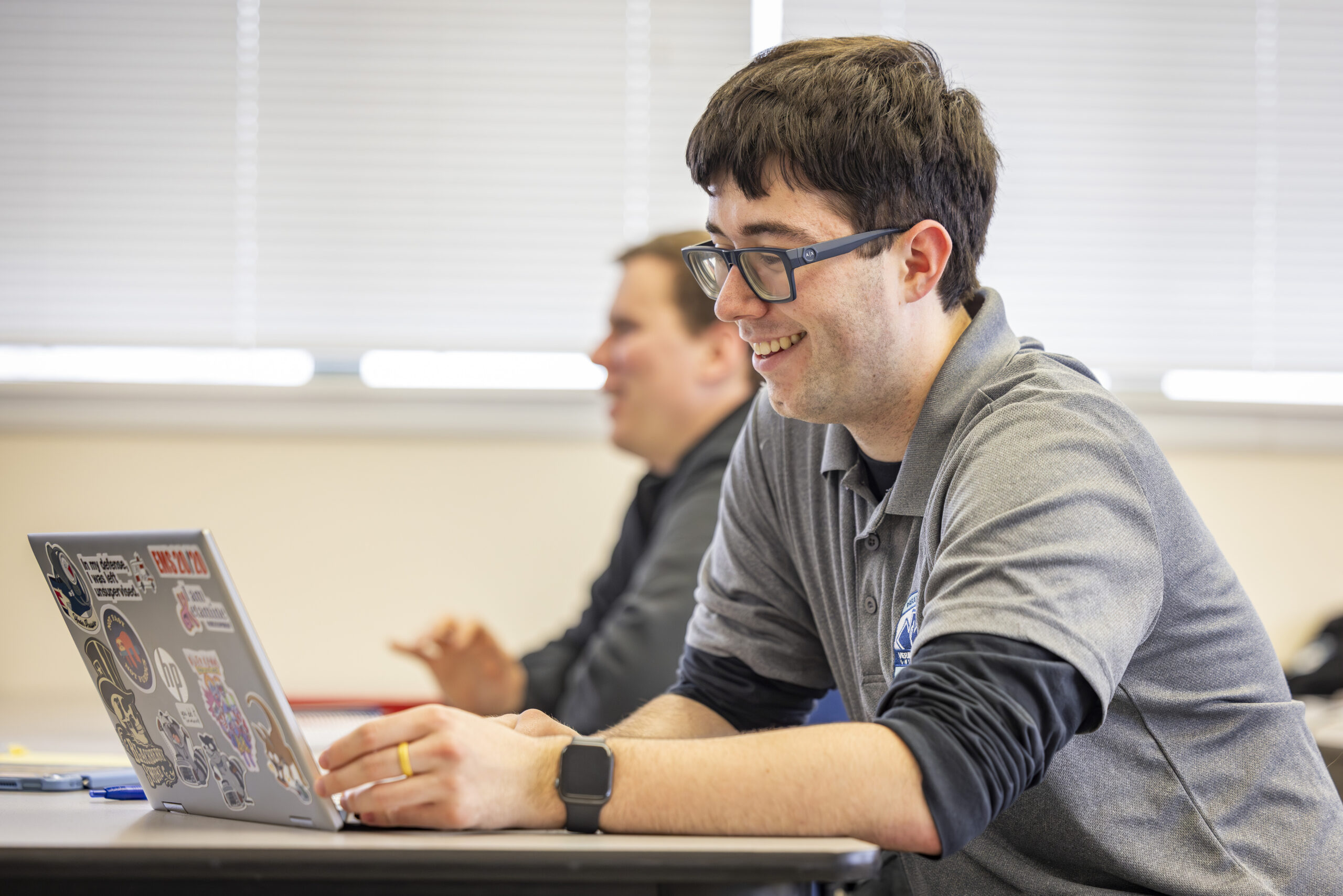 A man sitting at a table in a classroom, working on a laptop
