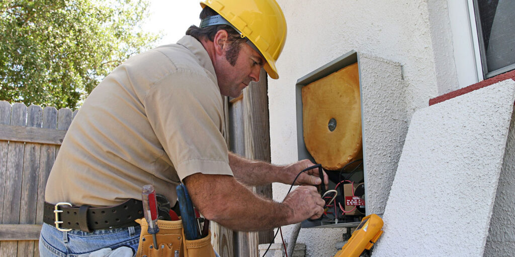 A man wearing a hard hat doing electrical work on an outdoor HVAC unit