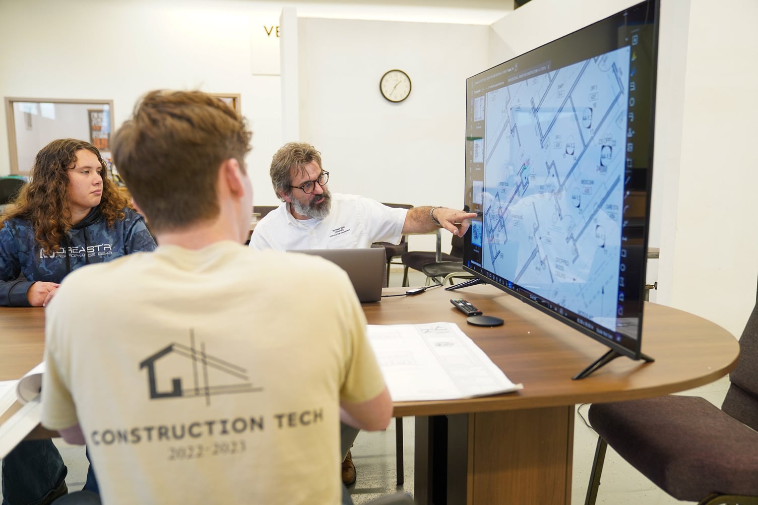 Three people sitting at table and inspecting a blueprint displayed on a large monitor